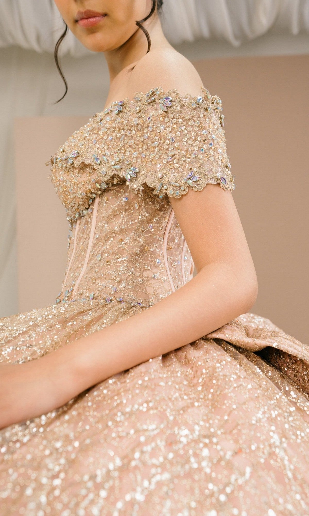 A person wearing the Quinceanera Dress 8600JQ By Cinderella Couture, an off-shoulder beige glittery gown featuring beaded embellishments and a corset bodice, sits against a neutral background.